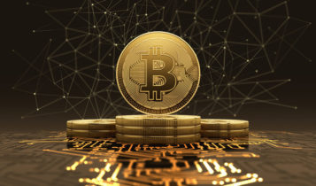 logo of a bitcoin on a golden coin that is standing on stacked coins as they stand on a motherboard