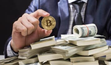 a business man holding a bitcoin coin and a pile of cash below the coin.